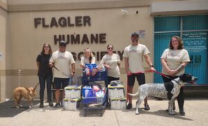 Six people stand outside the Flagler Humane Society with two dogs and a cart full of pet supplies They are all wearing matching beige "Alliance" shirts and smiling at the camera