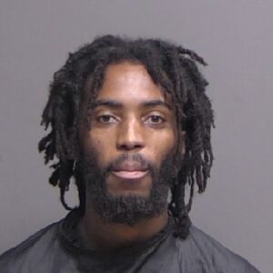 A man with medium length dreadlocks and a full beard looks directly at the camera in a neutral expression, wearing a dark garment against a plain gray background