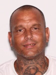 A middle aged man with a shaved head, visible tattoos on his neck, facial stubble, and gold teeth smiles slightly while wearing a white shirt and a chain necklace, against a plain light background