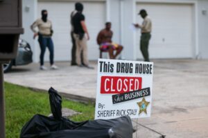 A sign reading "This drug house closed for business by Sheriff Rick Staly" is posted near a driveway, where masked officers and a seated, shirtless man are visible in front of garage doors