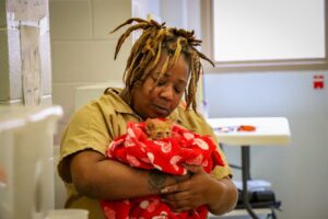 A person with dreadlocks wearing a tan uniform gently cradles a small orange kitten wrapped in a red blanket with white hearts, inside a brightly lit room