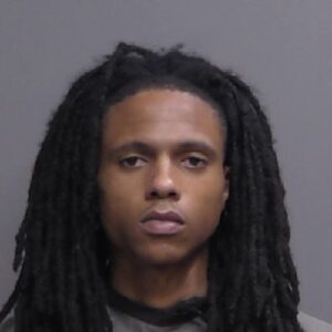 A young man with long, dark dreadlocks and a neutral expression is facing the camera against a plain gray background