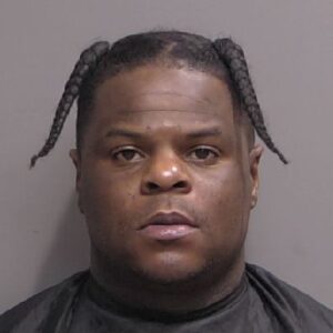 A man with two braids wears a dark cape and faces forward in a neutral, official photo against a plain background