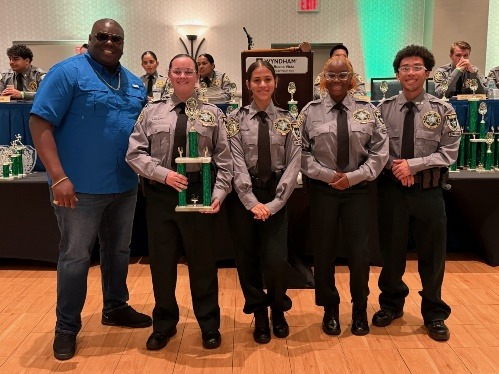 Five people pose indoors for a photo; four are in matching gray uniforms holding a trophy, and one is in a blue shirt and sunglasses Several trophies are displayed on tables in the background