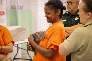 A woman in an orange uniform smiles while holding a gray kitten Two people in uniforms, one apparently a law enforcement officer, stand nearby in a brightly lit indoor setting