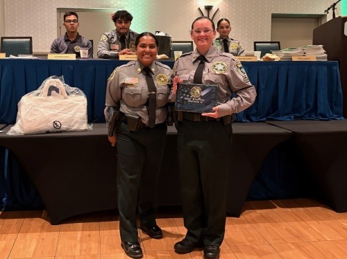 Two uniformed officers stand smiling, one holding a plaque, in front of a table with three seated individuals and awards on display The setting appears to be a formal ceremony or awards event
