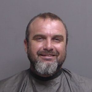 A middle aged man with short brown hair and a gray beard is smiling slightly He is wearing a dark cape or smock and is posed against a plain gray background