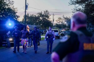 A shirtless man in colorful shorts is escorted by armed law enforcement officers in tactical gear at dusk, with police vehicles and flashing lights in the background An officer stands in the foreground