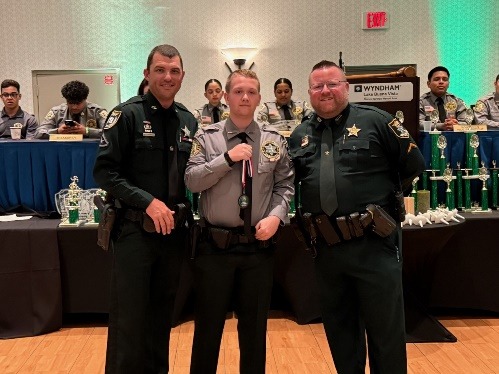 Three uniformed officers stand together indoors, smiling The center officer, holding a medal, is flanked by two others Behind them, more uniformed individuals sit at a table with green trophies displayed on stage