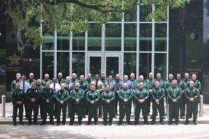 A large group of uniformed law enforcement officers stands in rows outside a building with large glass windows and doors, posing for a group photo under a leafy tree