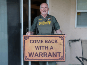A smiling sheriff in uniform stands outside a building, holding a large sign that reads "COME BACK WITH A WARRANT