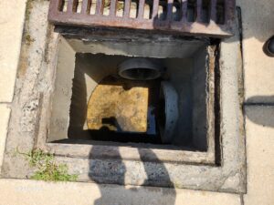 Open storm drain with a metal grate partially lifted, showing the concrete interior, a pipe, some water at the bottom, and shadows of people standing nearby Grass and pavement are visible at the edges