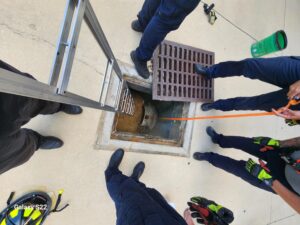 Five people in uniform stand around an open manhole with a metal ladder placed inside One person holds an orange rope, and safety equipment is visible nearby on the concrete ground