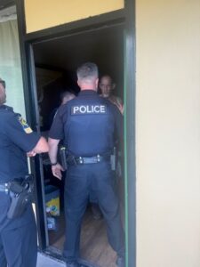 Three police officers stand at the doorway of a building, speaking with a man inside The officer in the center has "POLICE" written on the back of his vest The man inside appears to be engaging with the officers