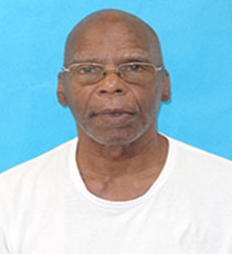 An older man with glasses and a bald head wearing a white t shirt is positioned in front of a solid light blue background He is looking directly at the camera with a neutral expression