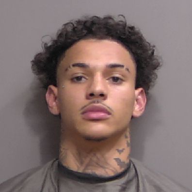 A young man with short curly hair, a thin mustache, and tattoos on his neck and face looks straight at the camera against a plain background He is wearing a dark colored garment