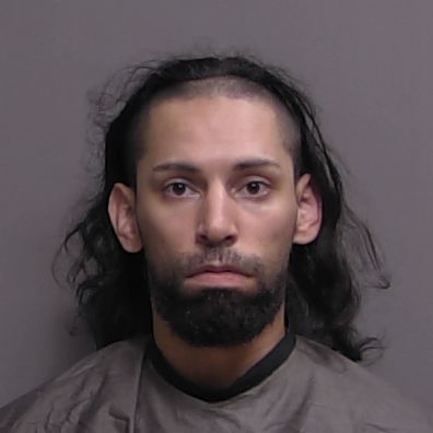 A person with long dark hair, a beard, and mustache, wearing a dark colored garment, looks directly at the camera against a plain, neutral background
