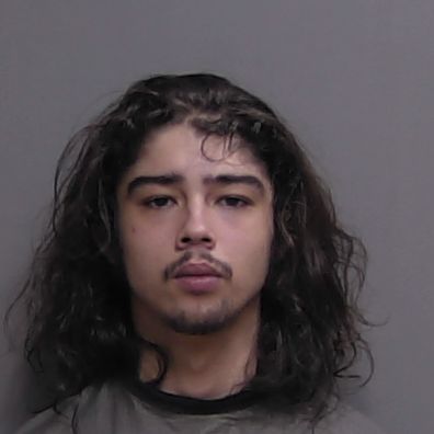 A young man with long, wavy dark hair and a mustache is facing the camera with a neutral expression, standing against a plain gray background