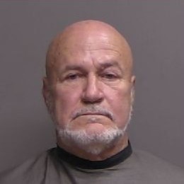 A bald older man with a white beard, wearing a dark cape or smock, stares straight at the camera against a plain gray background