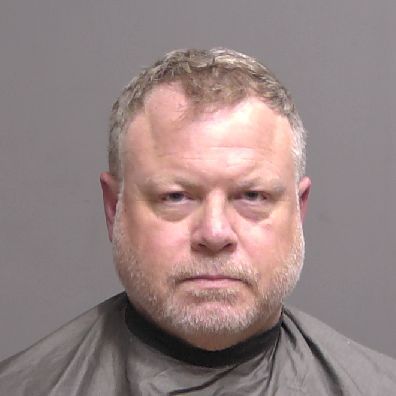 A middle aged man with short, light brown hair and a trimmed beard is wearing a dark drape, standing against a plain gray background, facing the camera with a neutral expression