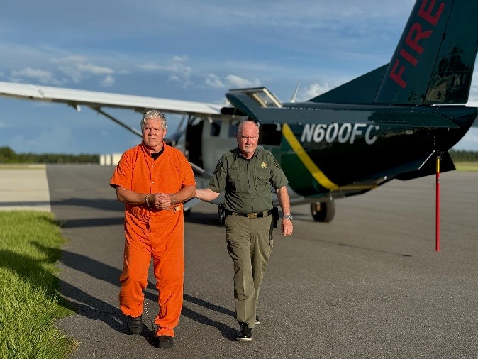 A man in an orange jumpsuit with handcuffs is escorted by a uniformed officer across an airstrip, with a small airplane marked "NFC" in the background