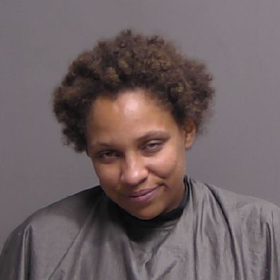A person with short curly hair wearing a gray drape is standing against a plain gray background, looking slightly downward with a subtle smile