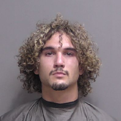 A young man with curly, shoulder length hair and a short goatee faces the camera for a mugshot He wears a dark drape or smock and stands against a plain, light background