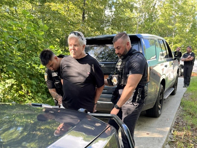 An older man in a black shirt is being handcuffed by two police officers beside a dark SUV on a wooded roadside, while more officers stand in the background