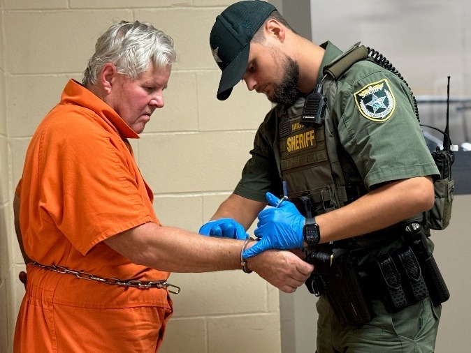 A law enforcement officer in a green sheriff's uniform handcuffs an older man in an orange prison jumpsuit and waist chains, inside a building with beige brick walls