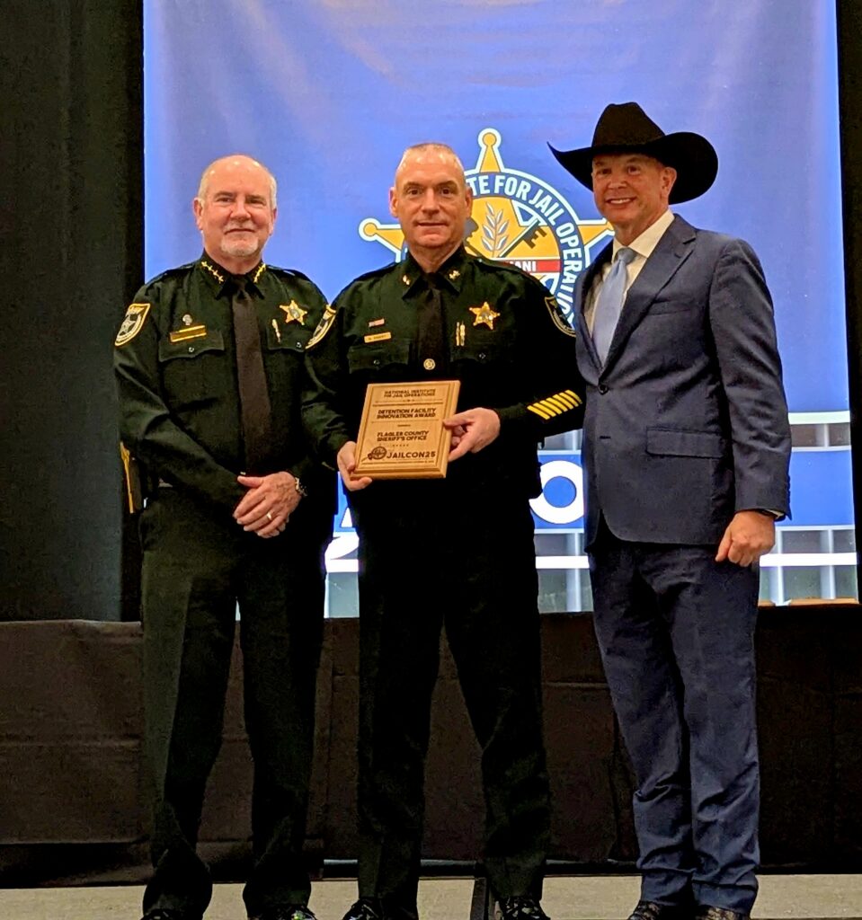 Three men stand together at an awards event Two are in sheriff uniforms and one is in a suit and cowboy hat The man in the center holds a plaque A blue banner with a sheriff’s badge logo is in the background Three men stand together at an awards event Two are in sheriff uniforms and one is in a suit and cowboy hat The man in the center holds a plaque A blue banner with a sheriff’s badge logo is in the background