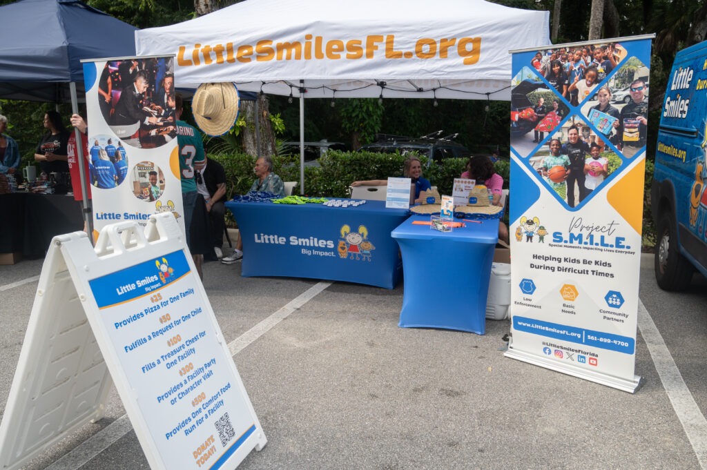 Outdoor booth for Little Smiles Florida with banners, an information board, and a table with volunteers and promotional items, promoting support for kids during difficult times Outdoor booth for Little Smiles Florida with banners, an information board, and a table with volunteers and promotional items, promoting support for kids during difficult times