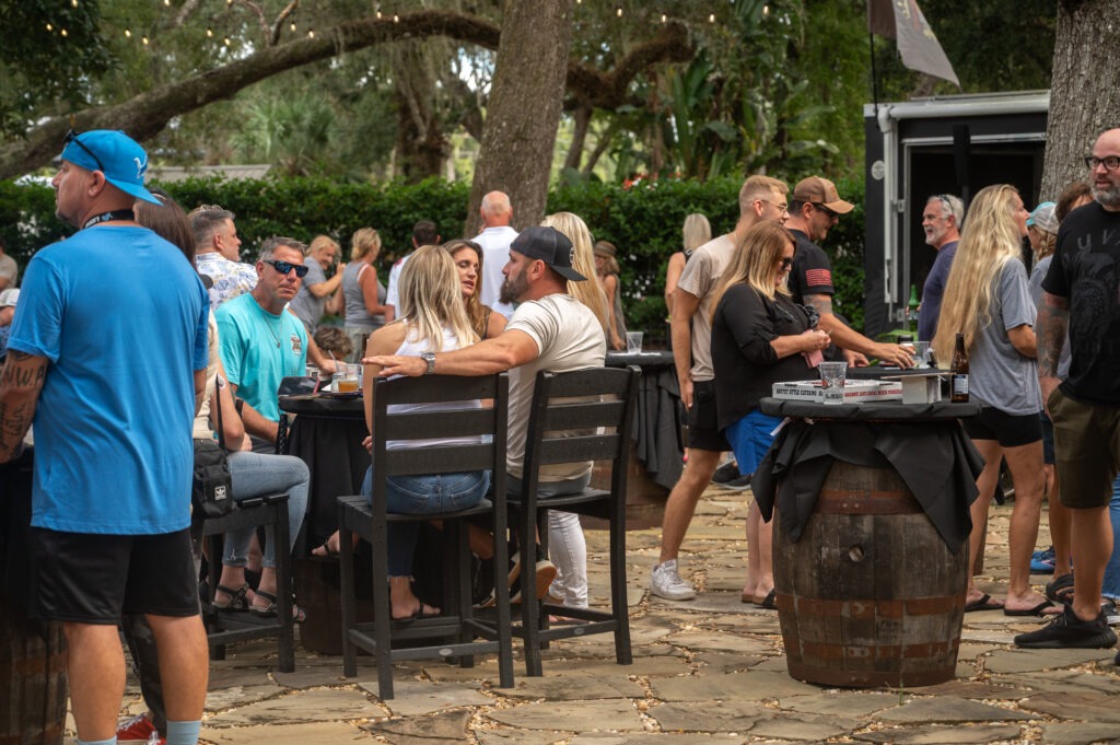 People gather outdoors at a casual event, sitting and standing around tables made from barrels Trees, string lights, and greenery create a relaxed atmosphere Most guests are chatting and enjoying food or drinks People gather outdoors at a casual event, sitting and standing around tables made from barrels Trees, string lights, and greenery create a relaxed atmosphere Most guests are chatting and enjoying food or drinks