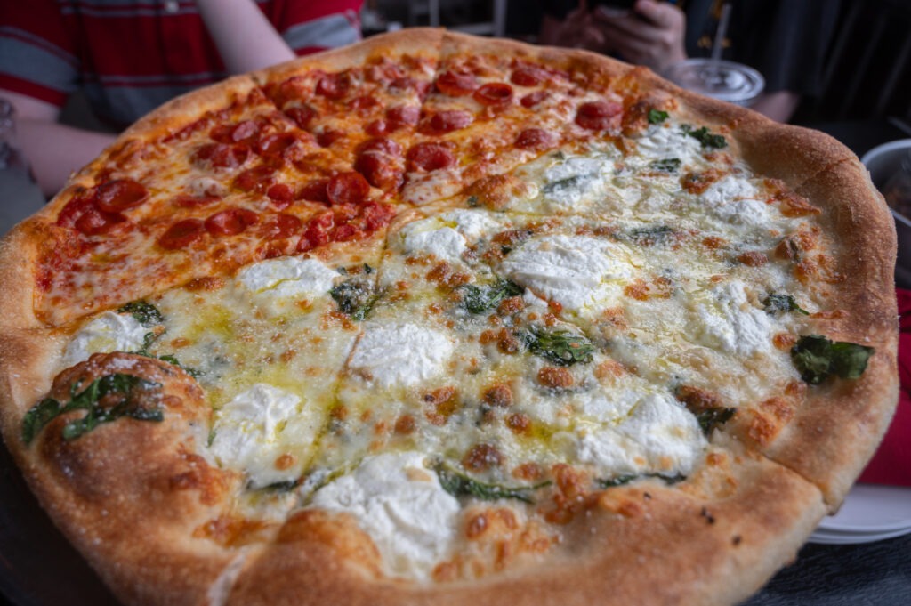 A large pizza with half topped with mini pepperoni slices and the other half topped with ricotta cheese and spinach The pizza sits on a table with drinks and people in the background A large pizza with half topped with mini pepperoni slices and the other half topped with ricotta cheese and spinach The pizza sits on a table with drinks and people in the background