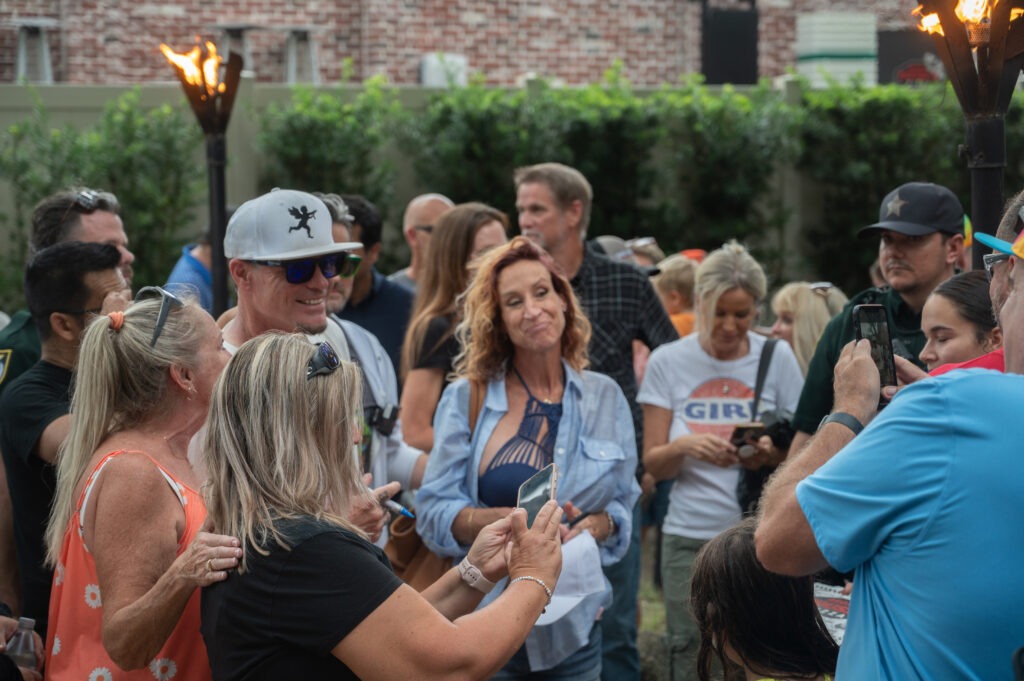 A group of people gather outdoors, some smiling and posing for photos A woman in the center holds a phone, while tiki torches burn nearby Trees and a brick wall are visible in the background A group of people gather outdoors, some smiling and posing for photos A woman in the center holds a phone, while tiki torches burn nearby Trees and a brick wall are visible in the background