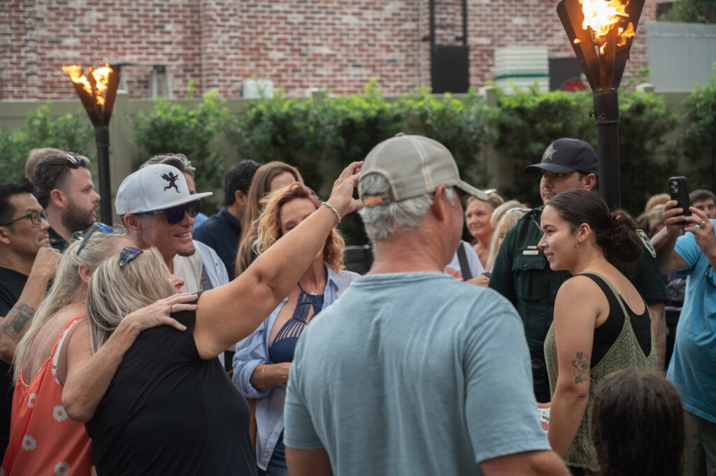 A group of people gathers outdoors near tiki torches, some smiling and posing for photos A woman in front is talking to the group while others take pictures Trees and a brick building are in the background A group of people gathers outdoors near tiki torches, some smiling and posing for photos A woman in front is talking to the group while others take pictures Trees and a brick building are in the background