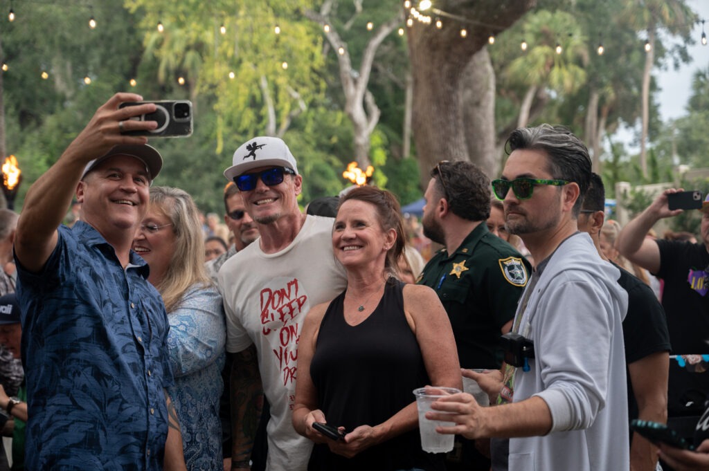 A group of people smiles and poses for a selfie at an outdoor event, with string lights and trees in the background One man holds a phone, and others wear sunglasses and casual clothing A police officer stands among them A group of people smiles and poses for a selfie at an outdoor event, with string lights and trees in the background One man holds a phone, and others wear sunglasses and casual clothing A police officer stands among them