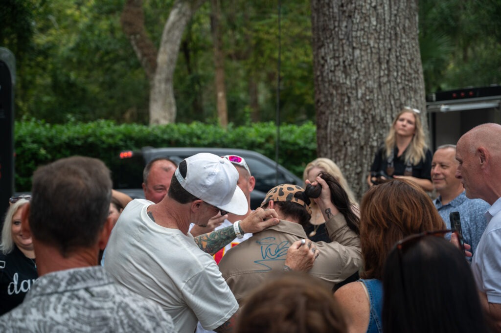 A man in a white cap signs the back of someone's shirt with a blue marker while a group of people watch outdoors, surrounded by trees and greenery A man in a white cap signs the back of someone's shirt with a blue marker while a group of people watch outdoors, surrounded by trees and greenery