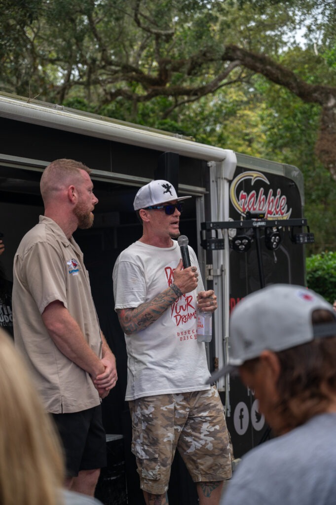Two men stand and speak in front of a trailer with the "Crappie" logo; one holds a microphone and paper Trees are in the background, and an audience member wearing a cap is in the foreground Two men stand and speak in front of a trailer with the "Crappie" logo; one holds a microphone and paper Trees are in the background, and an audience member wearing a cap is in the foreground