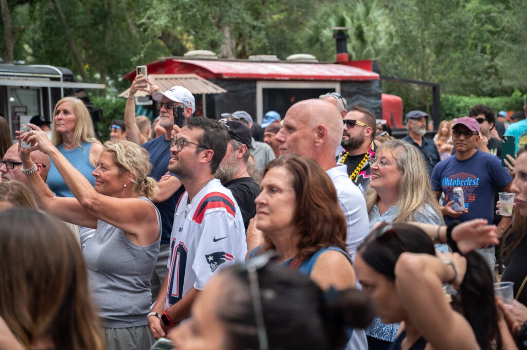 A diverse crowd of people, some smiling and taking photos, gather outdoors at an event near food trucks and trees on a sunny day A diverse crowd of people, some smiling and taking photos, gather outdoors at an event near food trucks and trees on a sunny day