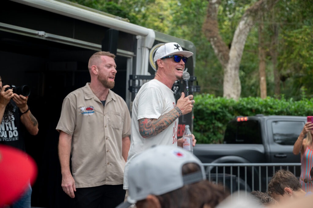 A man in a white t shirt, sunglasses, and a cap speaks into a microphone outdoors, while another man in a beige shirt stands beside him A crowd watches, and trees and a black truck are in the background A man in a white t shirt, sunglasses, and a cap speaks into a microphone outdoors, while another man in a beige shirt stands beside him A crowd watches, and trees and a black truck are in the background