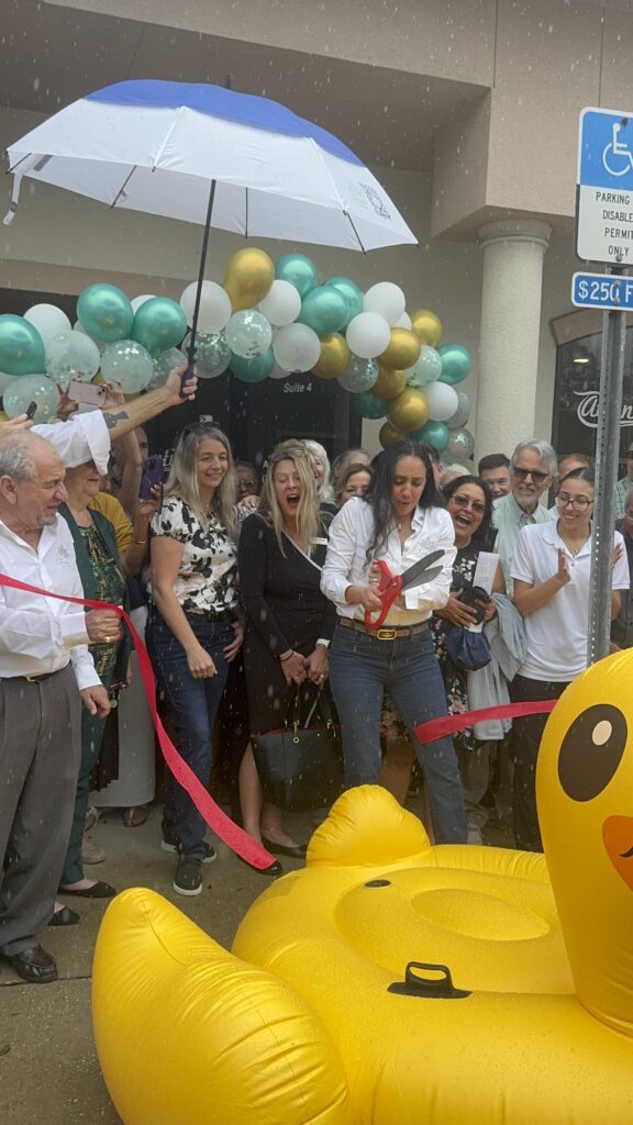 A woman cuts a red ribbon at an outdoor event, surrounded by smiling people, balloons, a large rubber duck float, and someone holding an umbrella A blue handicapped parking sign is visible on the right
