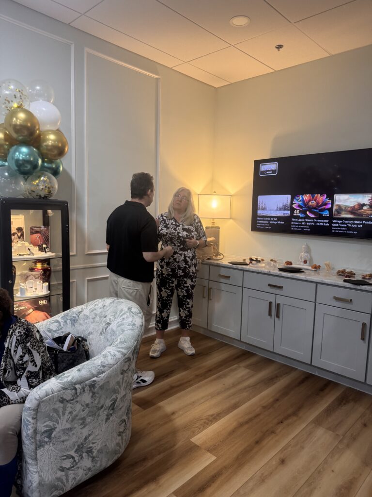 Two people stand and talk in a cozy room with balloons, a TV displaying various images, and a countertop with snacks Light wood flooring, white cabinets, and a soft armchair create a relaxed atmosphere Two people stand and talk in a cozy room with balloons, a TV displaying various images, and a countertop with snacks Light wood flooring, white cabinets, and a soft armchair create a relaxed atmosphere