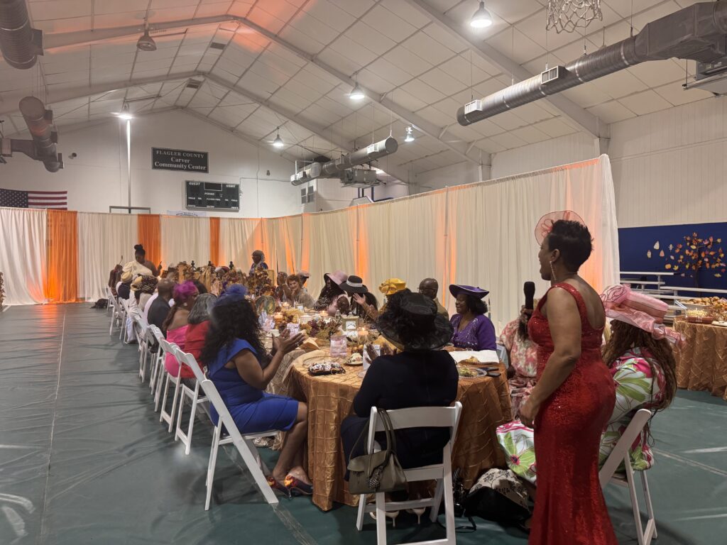 A large group of women in colorful dresses and hats are seated at long, decorated tables inside a gymnasium for a formal event One woman in a red dress stands and speaks, while others eat and converse A large group of women in colorful dresses and hats are seated at long, decorated tables inside a gymnasium for a formal event One woman in a red dress stands and speaks, while others eat and converse