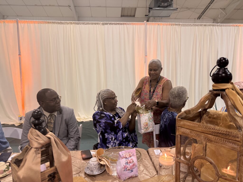 Four people sit at a decorated table with teacups and a lantern, while a woman standing hands a floral gift bag to an older woman with braided hair The background features cream colored curtains and warm lighting