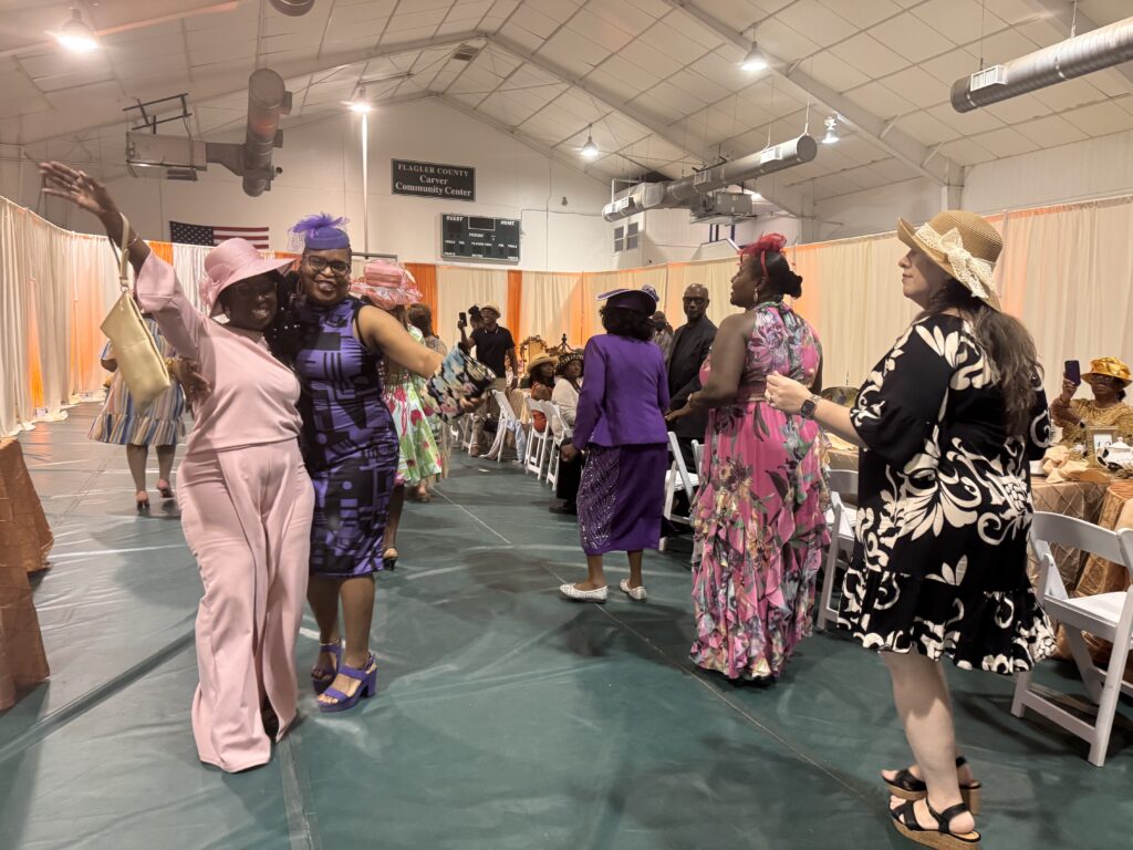 Women in colorful dresses and hats smile and pose for photos at a lively indoor gathering Two women in the foreground embrace and wave while others chat and mingle in the background The room features draped curtains and high ceilings