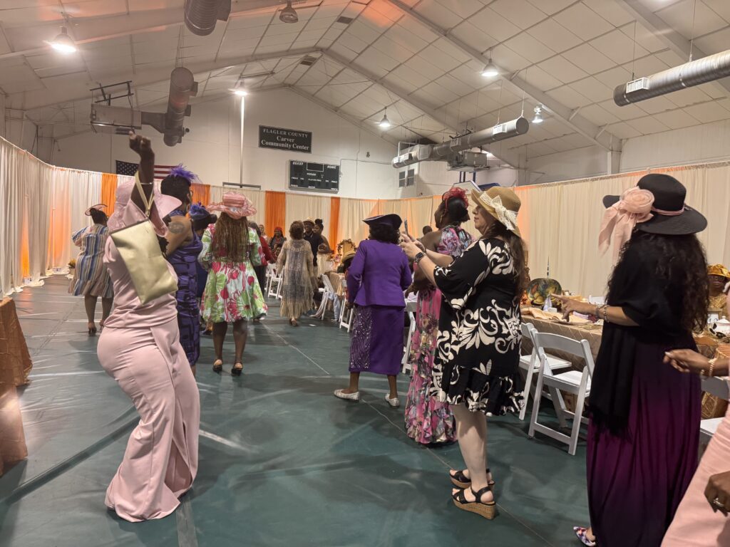 Women in colorful dresses and hats dance and mingle in a decorated indoor venue with draped curtains, tables, and chairs, suggesting a festive or formal event Women in colorful dresses and hats dance and mingle in a decorated indoor venue with draped curtains, tables, and chairs, suggesting a festive or formal event