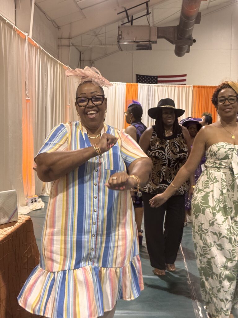 A smiling woman in a colorful striped dress dances in the foreground, while other women, some wearing hats and patterned outfits, dance behind her The scene takes place indoors with curtains and an American flag in the background A smiling woman in a colorful striped dress dances in the foreground, while other women, some wearing hats and patterned outfits, dance behind her The scene takes place indoors with curtains and an American flag in the background