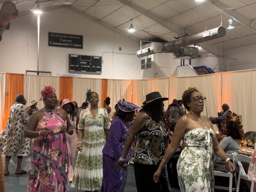 A group of women in colorful dresses and hats dance together inside the Flagler County Carver Community Center, decorated with orange and white curtains Other guests are seated in the background