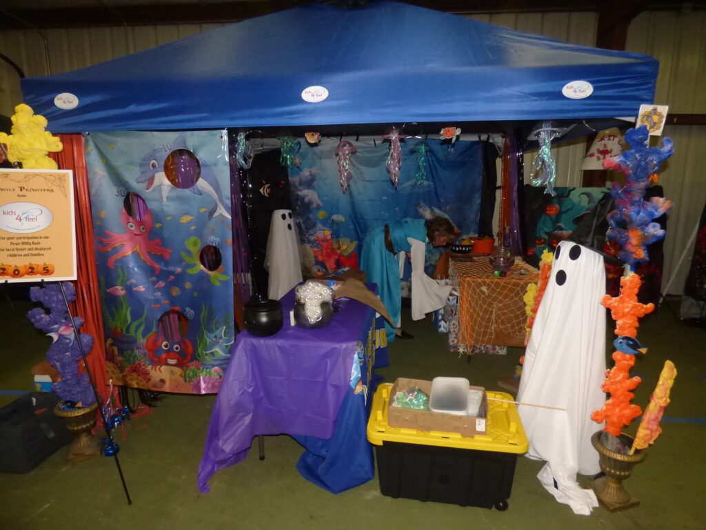 A colorful, undersea themed booth decorated with sea creature curtains, plastic coral, and Halloween ghost figures A blue canopy covers the setup, with tables holding candy bowls and festive decorations A colorful, undersea themed booth decorated with sea creature curtains, plastic coral, and Halloween ghost figures A blue canopy covers the setup, with tables holding candy bowls and festive decorations