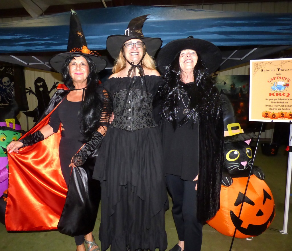 Three women dressed as witches in black costumes and hats pose and smile at a Halloween themed event, with festive decorations and a sign for a BBQ in the background Three women dressed as witches in black costumes and hats pose and smile at a Halloween themed event, with festive decorations and a sign for a BBQ in the background